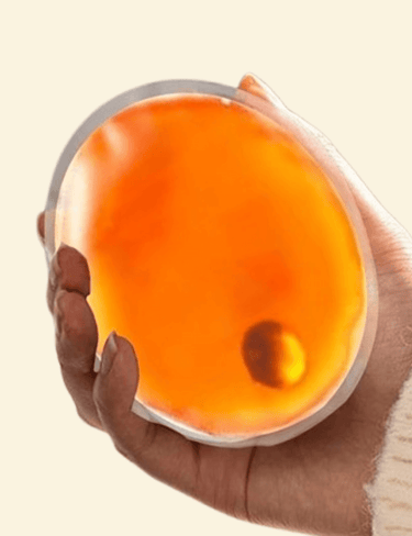 A close-up of a hand holding a bright orange reusable hand warmer against a light cream background.